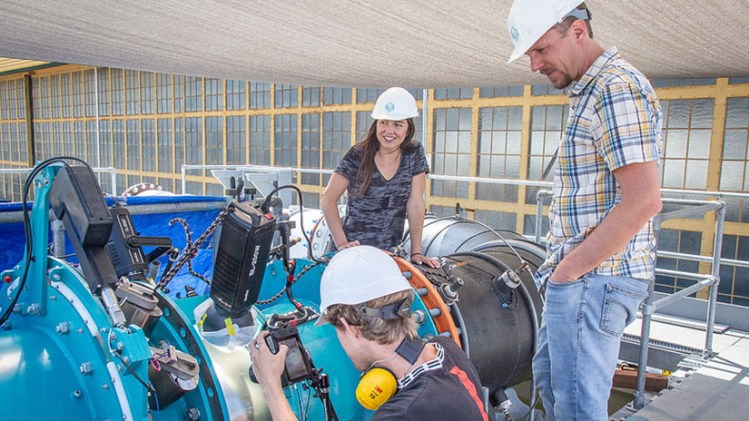 Three people in hard hats work around a large metal pipe.