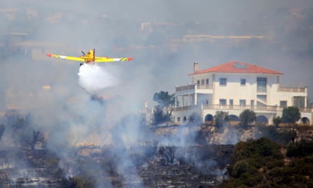 A yellow aircraft dropping water as it flies through thick smoke in front of a white-walled, red-roofed villa surrounded by trees