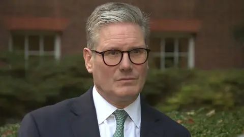 BBC The Prime Minister Sir Keir Starmer in a dark suit an white shirt with a green tie, wearing black-framed glasses, standing in the Downing Street garden 