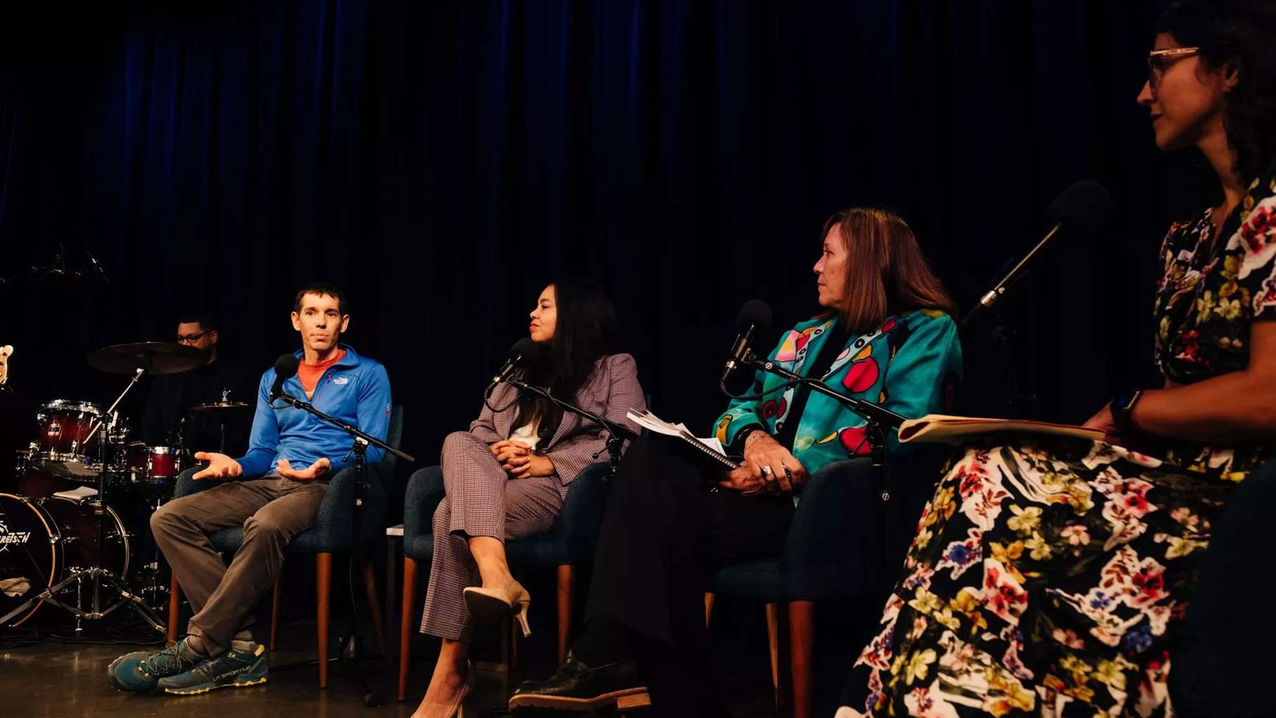 Five people are seated on a dark stage. At the far left, a person sits in shadow behind a drum set. Next is a person with short dark hair wearing an orange T-shirt and blue half-zip pullover; next is a person with long dark hair wearing a gray suit, followed by a person with medium-length brown hair wearing a teal jacket with colorful patches and holding papers on their lap. Closest to the camera is a person with patterned eyeglasses and wearing a flowered dress.