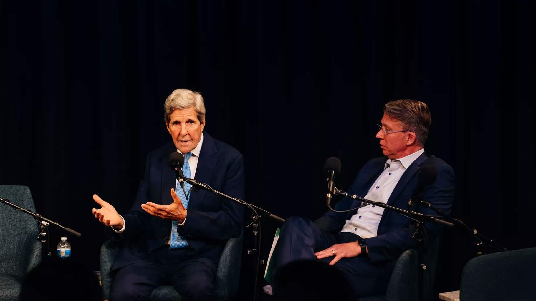 Two people sit in chairs on a stage with microphones in front of them. The person on the left, wearing a dark suit and blue tie, gestures as the person on the right leans back in the chair listening to the remarks.  