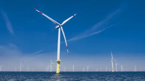 Getty Images A massive wind turbine in the middle of the sea, photographed on a sunny day with clear blue skies. In the background you can see around 30 more, stretching into the horizon.