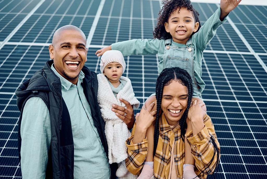 A smiling Black family standing in front of a large solar panel. The mother carries her daughter on her shoulders while the father carries their infant in his arms.