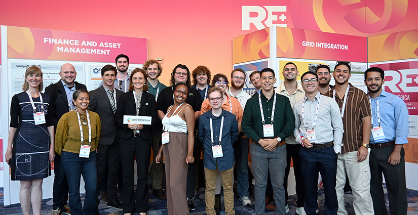 A group of 20 people stands smiling for the camera. There are research posters in the background and the logo for RE+.