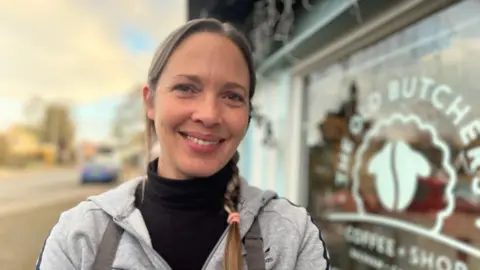 Emma Baugh/BBC Holly Venn is wearing an apron and grey jumper, she is smiling at the camera, in the background is the glass front of her coffee shop.