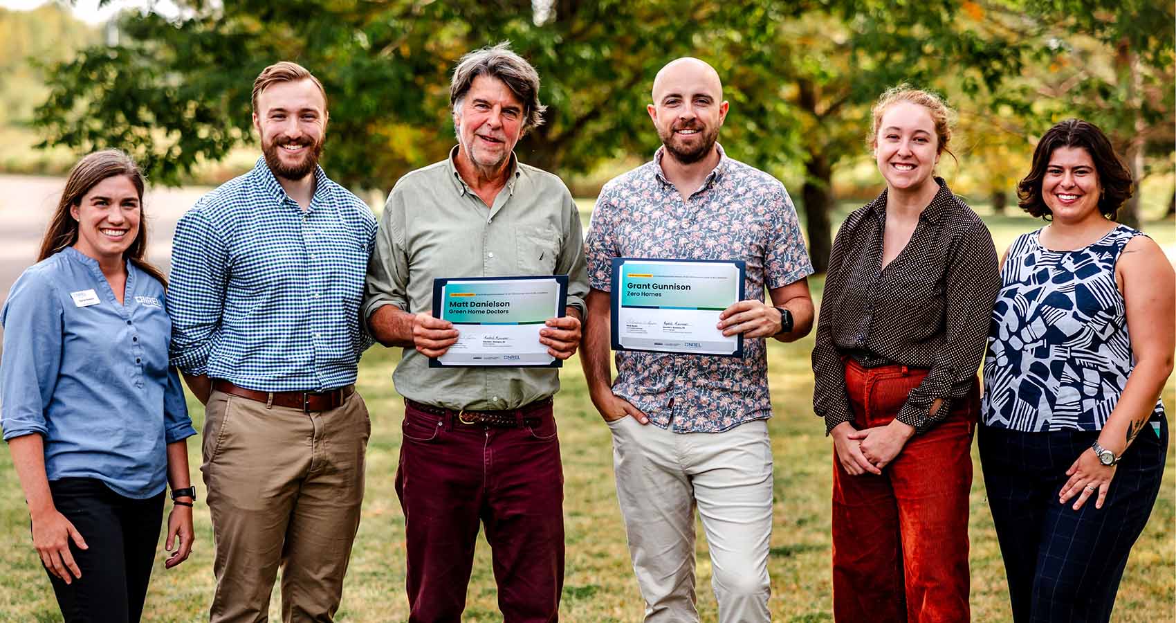 Six people stand side-by-side before a tree, two of which are holding certificates of completion for B-Biz. 