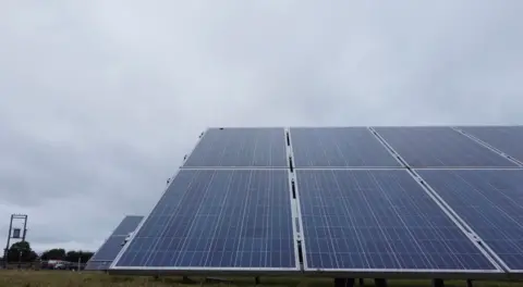 Solar panels in a field - the sky behind then is clouded over and an electrical transformer can be seen in the background.