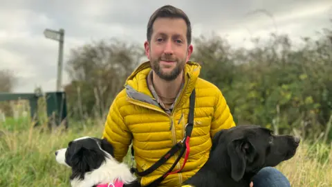 Emma Baugh/BBC Mike Davey is holding his two dogs, and wearing a yellow puffer jacket. He is on a walk on a countryside footpath.