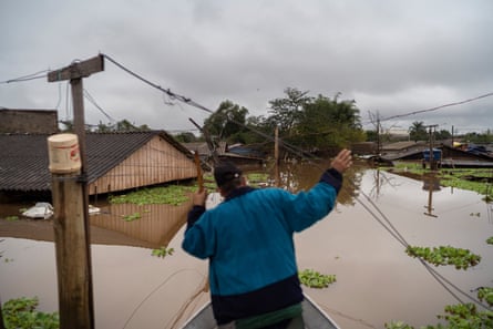 A man seen from behind as he uses a pole to steer his small boat towards buildings where water is almost up to the eaves 