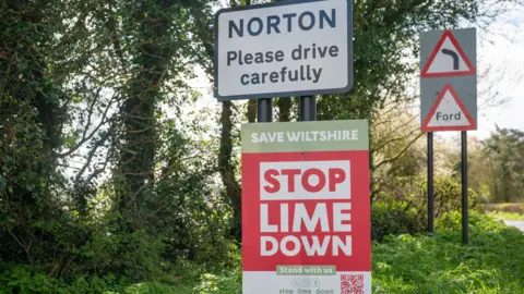 PA Media A village road sign for Norton with a temporary sign attached to the poles holding it up saying 'Stop Lime Down'. One the side of green verge with trees.