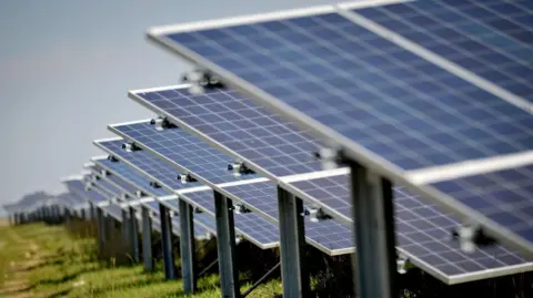 PA Media Rows of black and white solar panels on metal struts in a field.