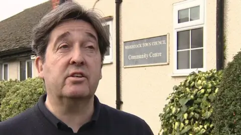 Mark Owen-Lloyd has short brown hair and is wearing a black knitted jumper. Behind him is a cream wall.